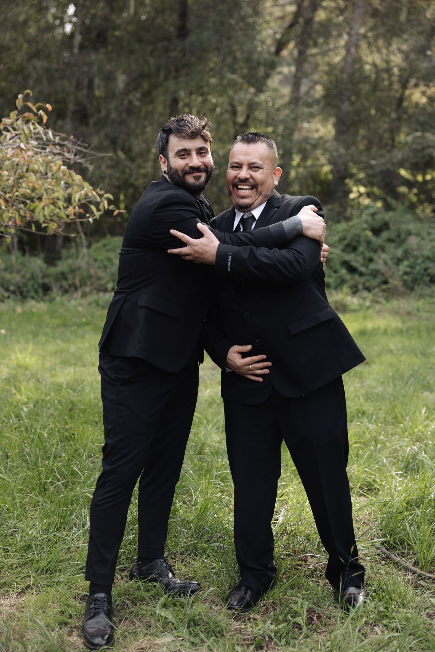 Two people in black suits share playful moments in a grassy outdoor setting.
