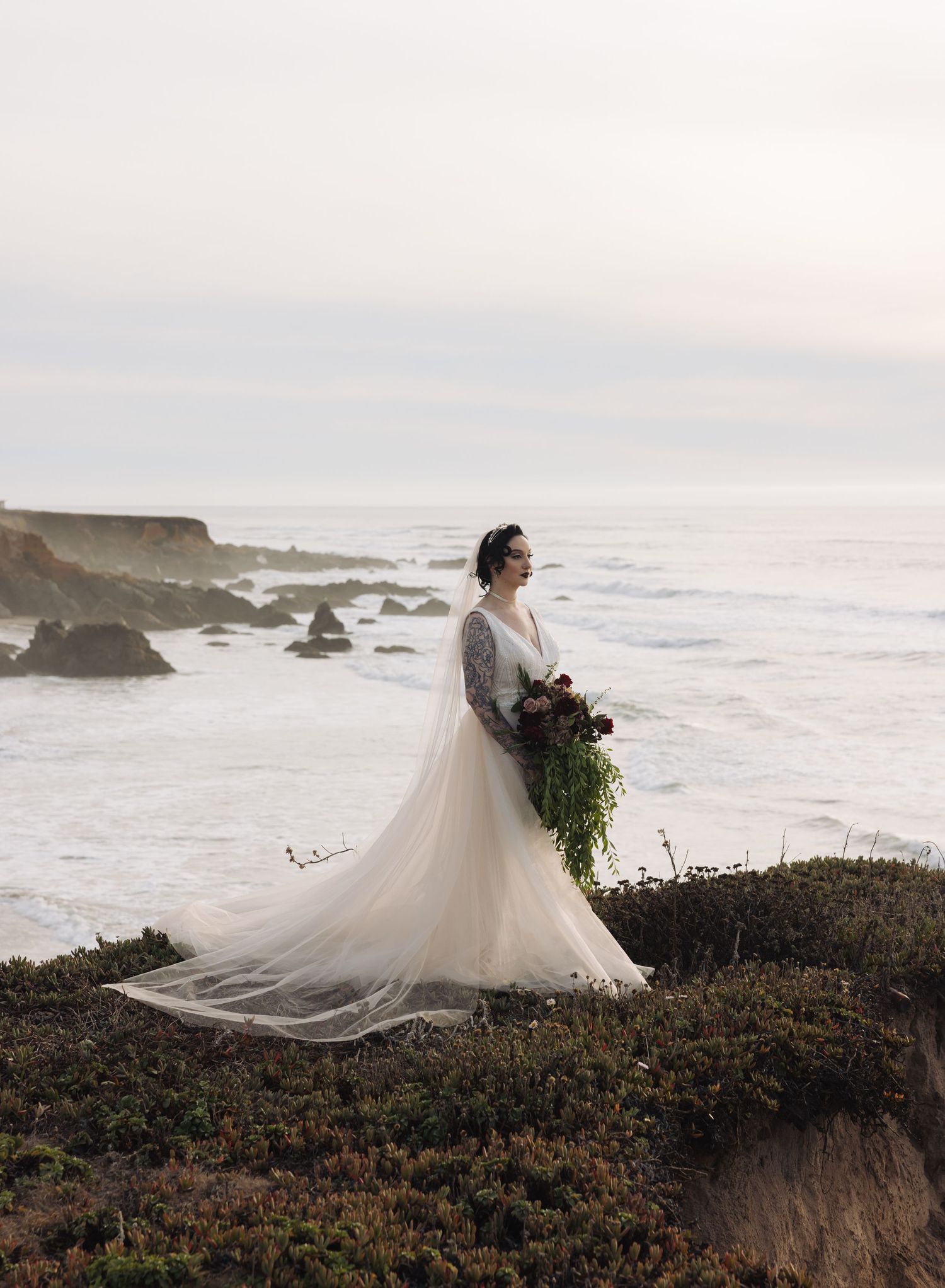 A bride in a flowing white dress stands on a coastal cliff overlooking the Pacific Ocean.