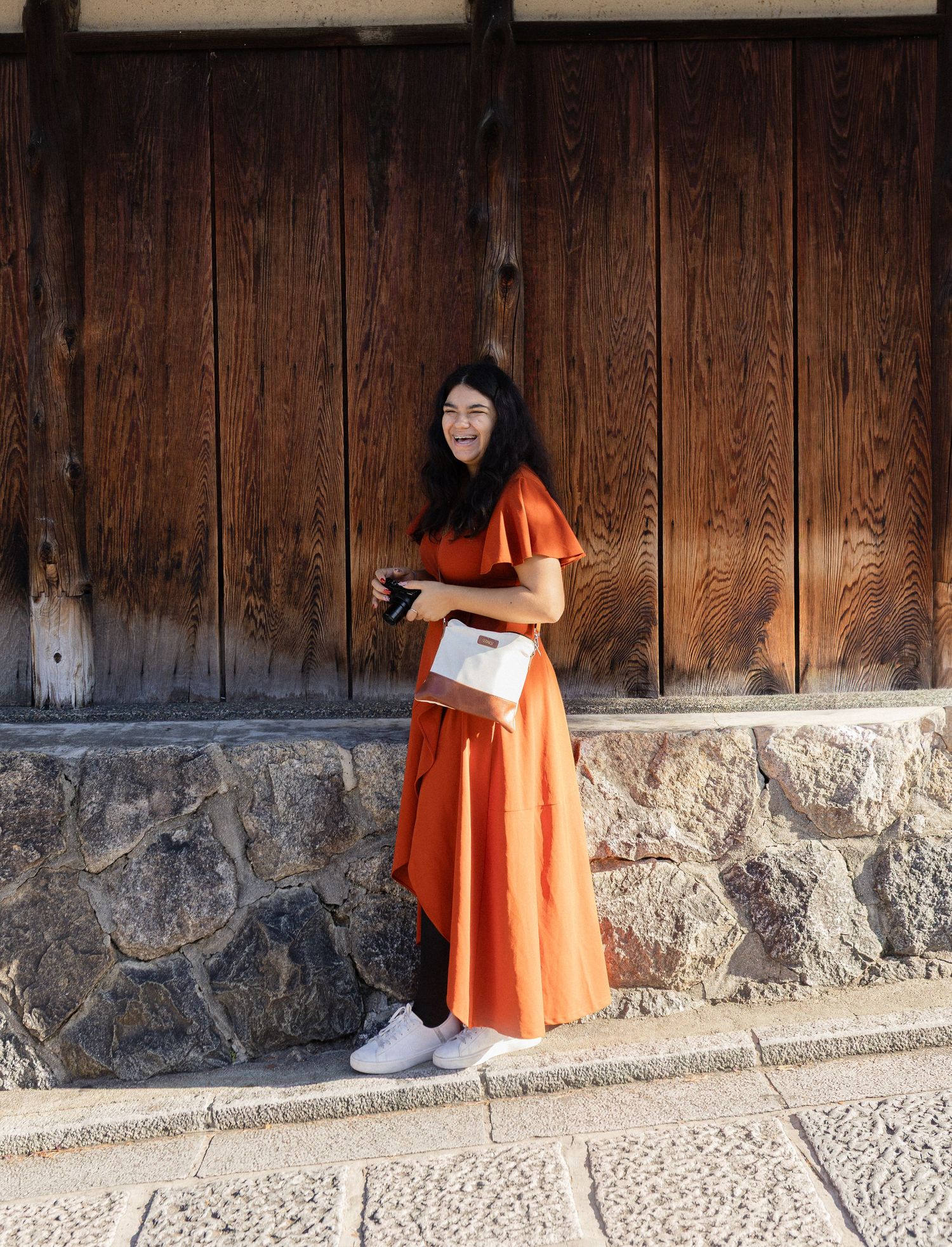Person in vibrant orange dress standing against rustic wooden wall with stone foundation.