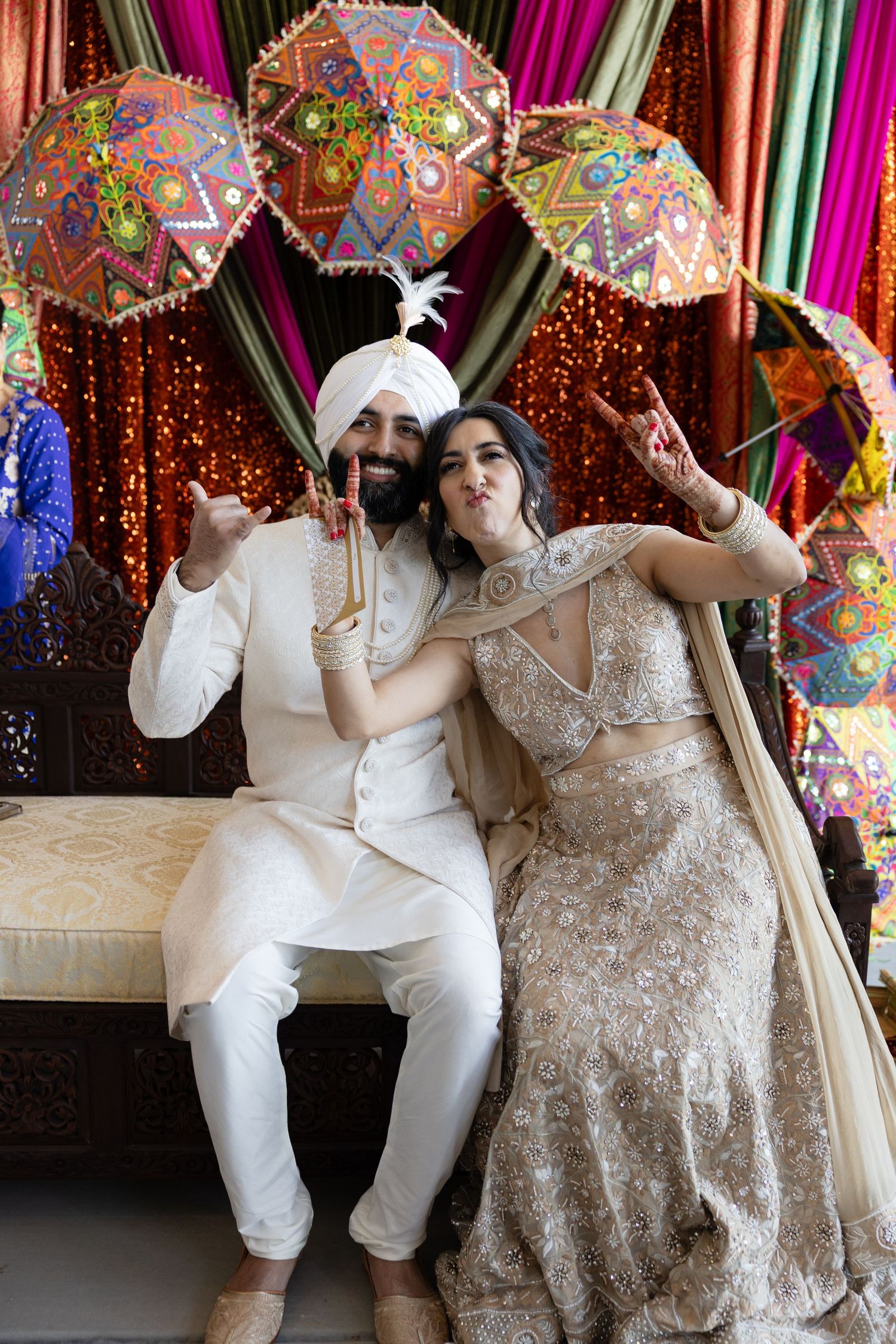 A couple poses joyfully under decorative umbrellas and colorful drapery at an Indian wedding celebration.
