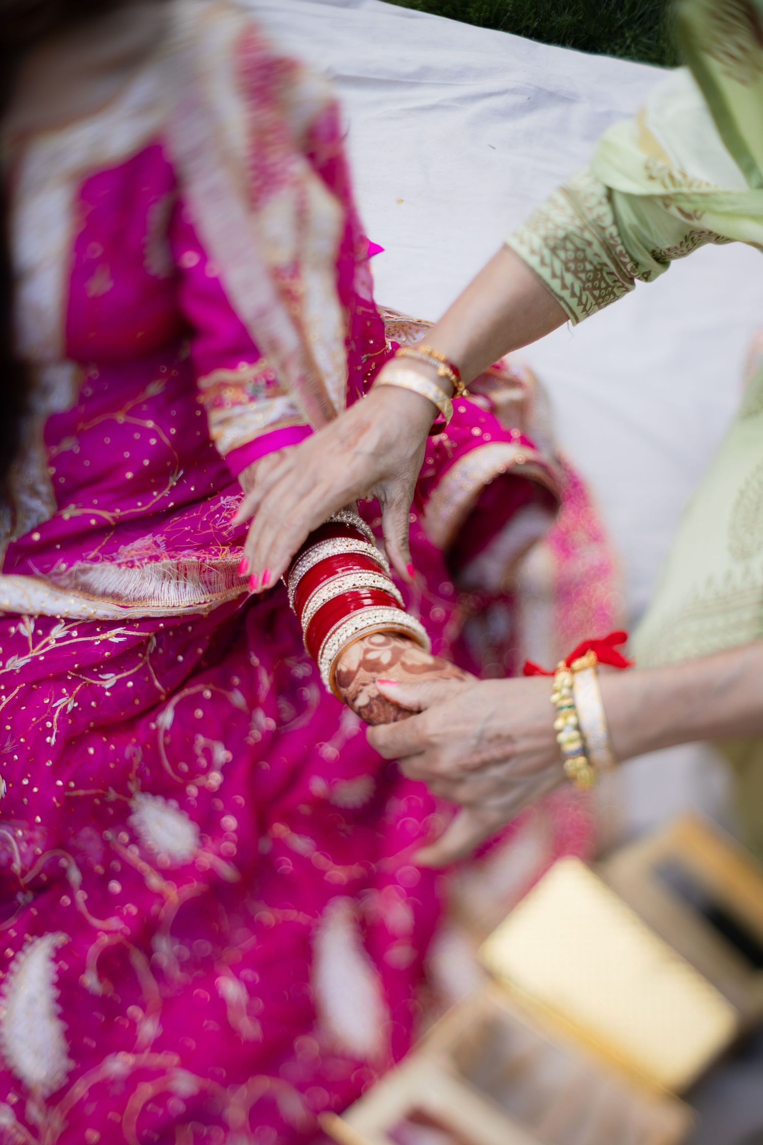 Traditional Indian wedding ceremony showing hands adorned with red bangles and gold jewelry against magenta fabric.