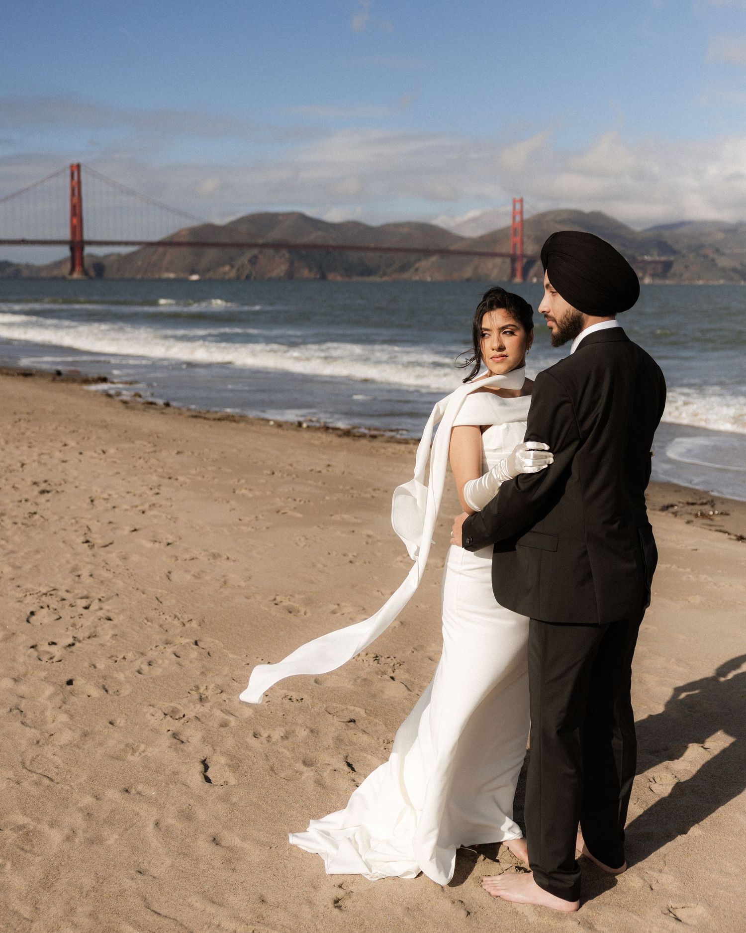 A couple in formal attire poses together on a sandy beach with the Golden Gate Bridge visible in the background.