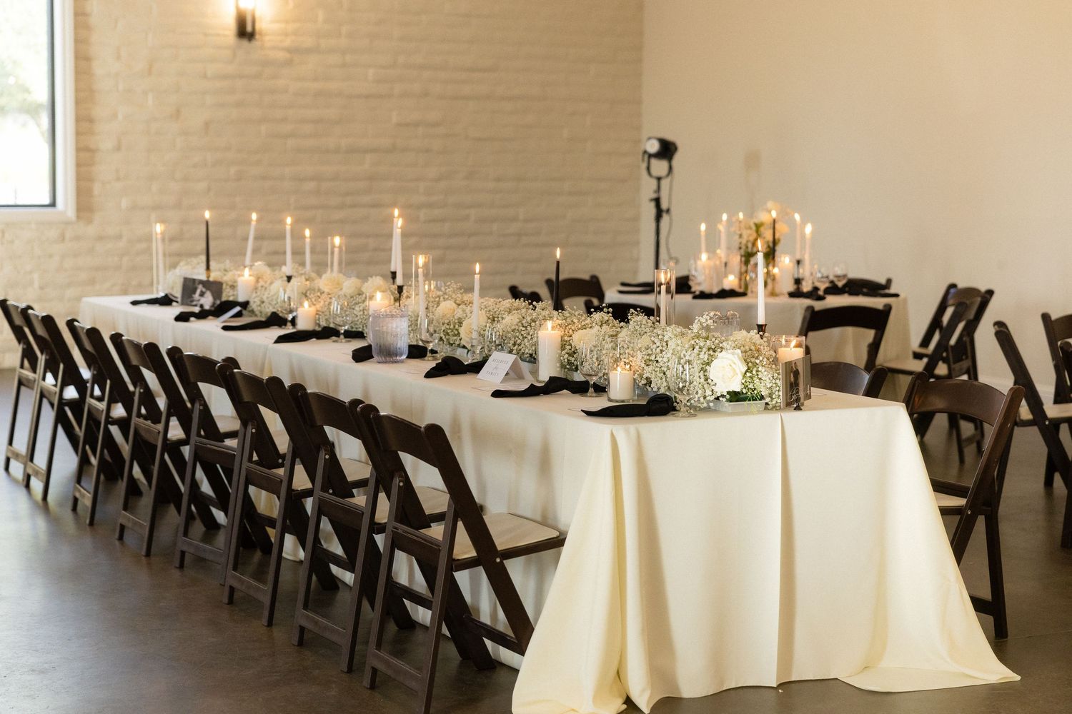 Elegant long banquet table with cream linens, candles, and floral centerpieces in a white brick venue.