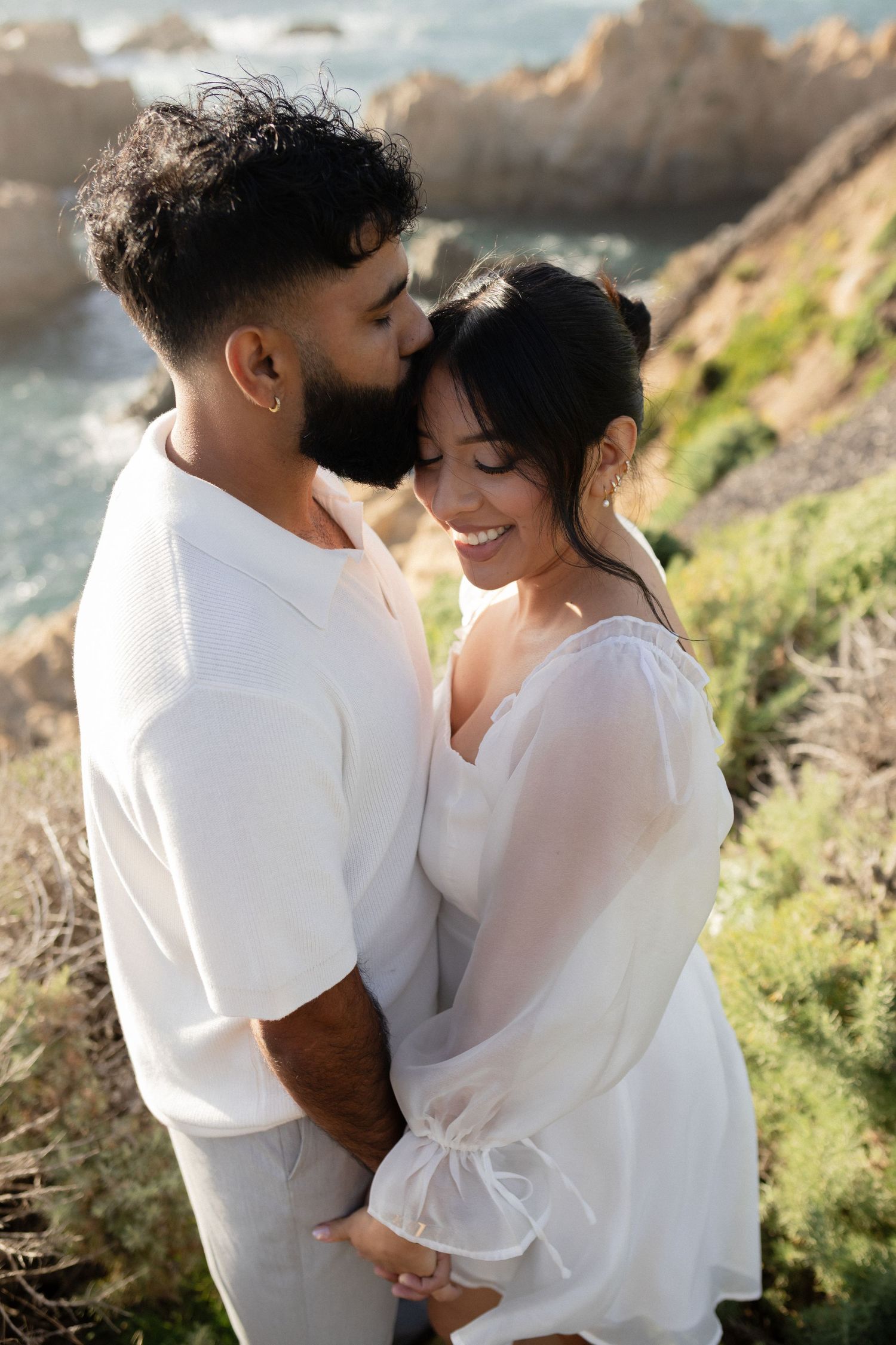 Couple in white outfits embracing on a coastal cliff, man kissing woman's temple as she smiles, ocean backdrop.