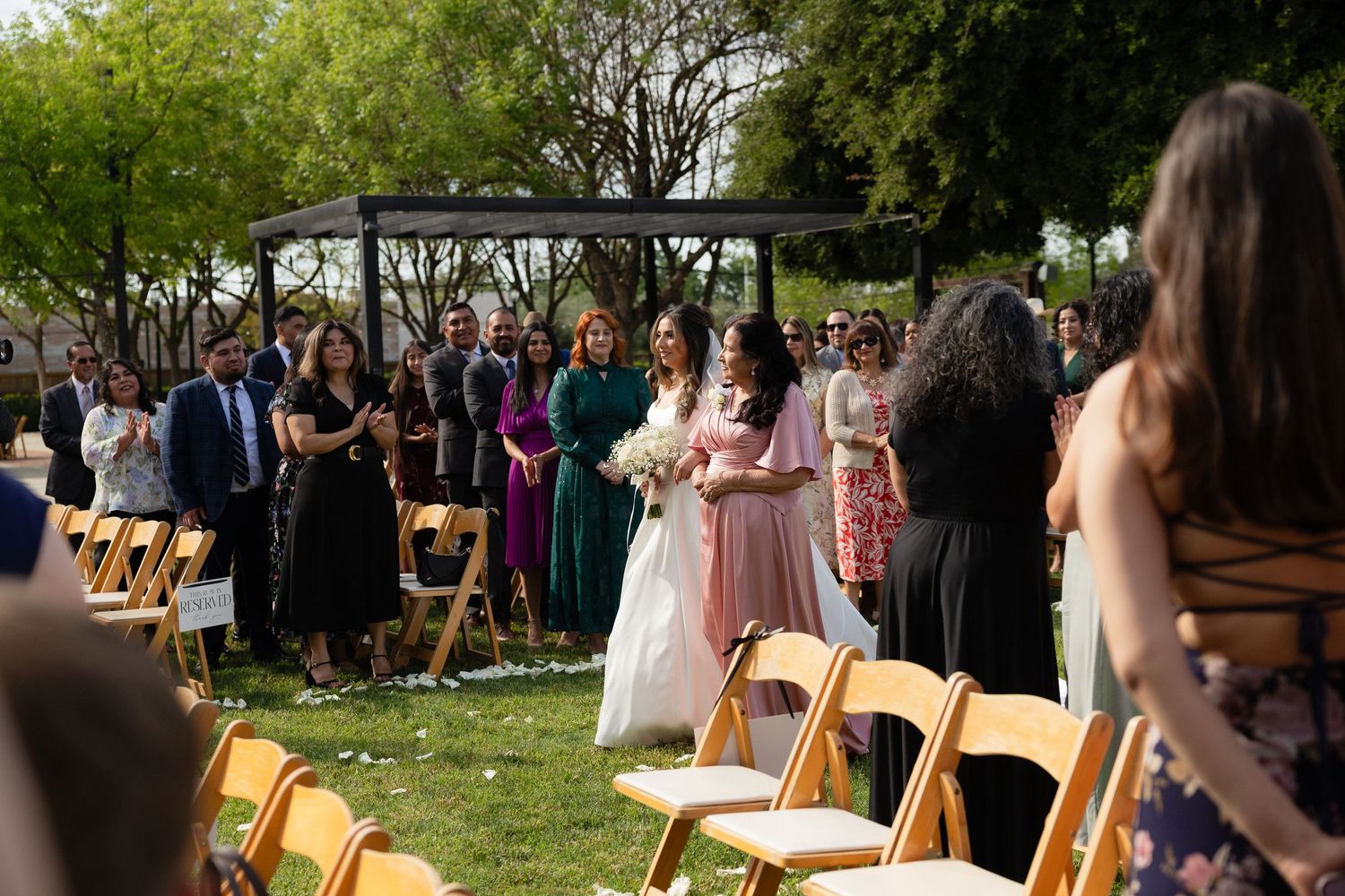 Bride walks down outdoor aisle with wedding party and guests at garden ceremony.