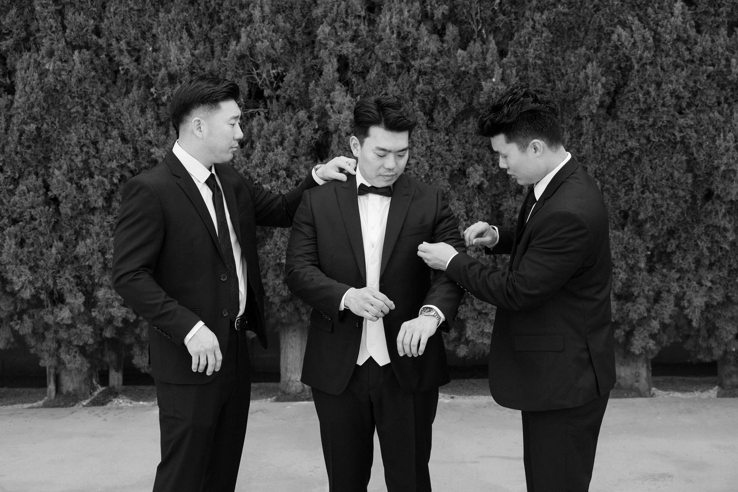 Three groomsmen in black tuxedos help each other prepare before a wedding ceremony outdoors.
