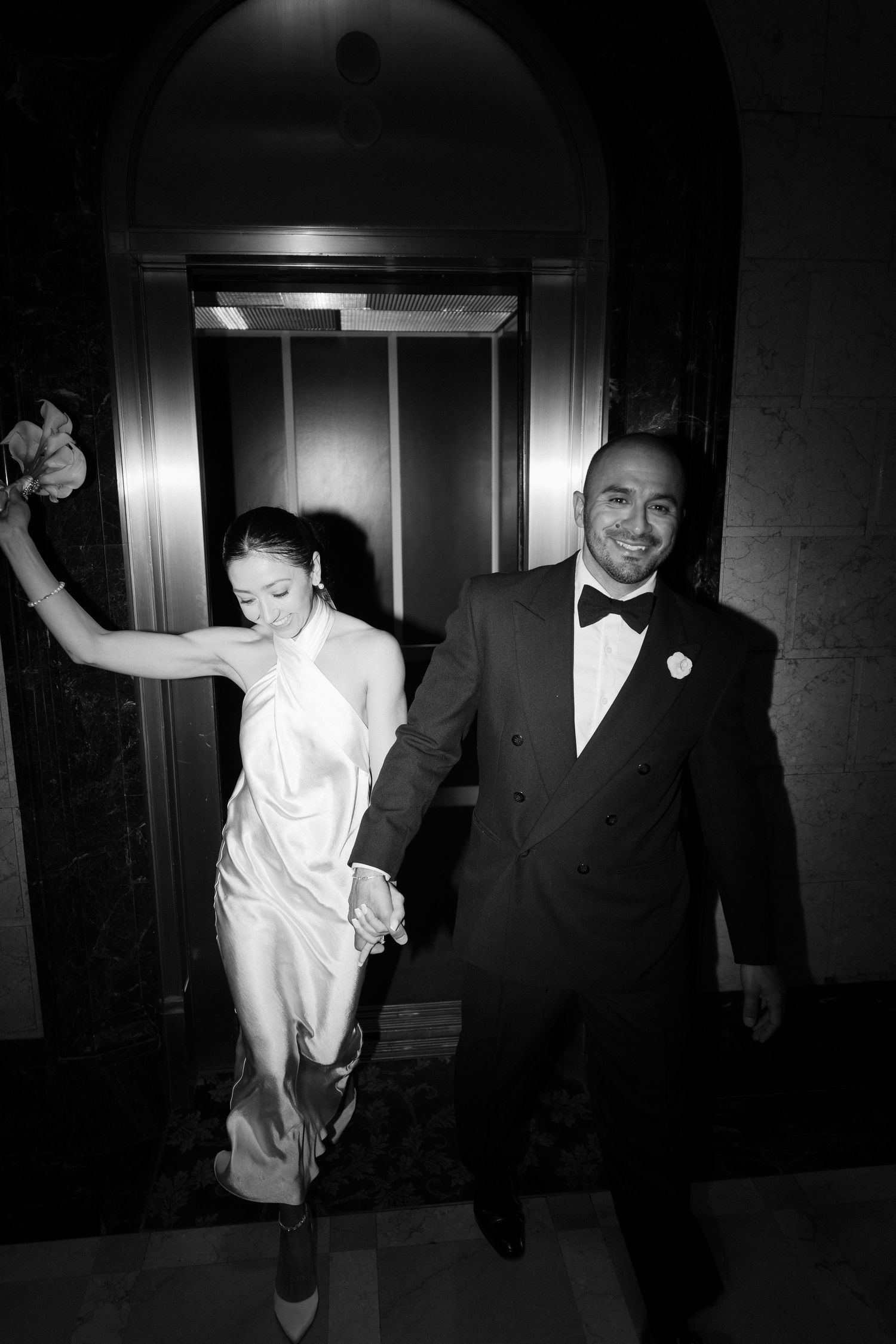 A joyful couple exits an elevator in formal attire, with the bride in white and groom in a black tuxedo and bowtie.