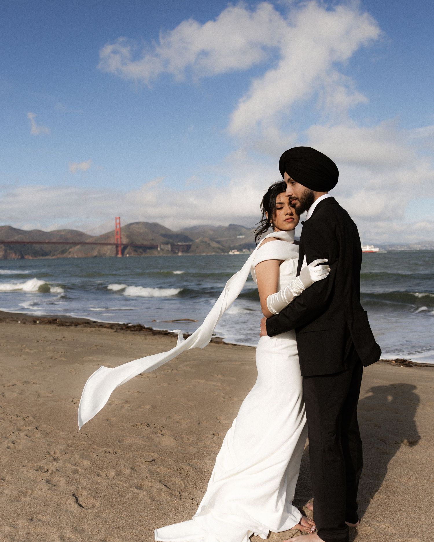 A romantic wedding couple embraces on a sandy beach with the Golden Gate Bridge and San Francisco Bay in the background.