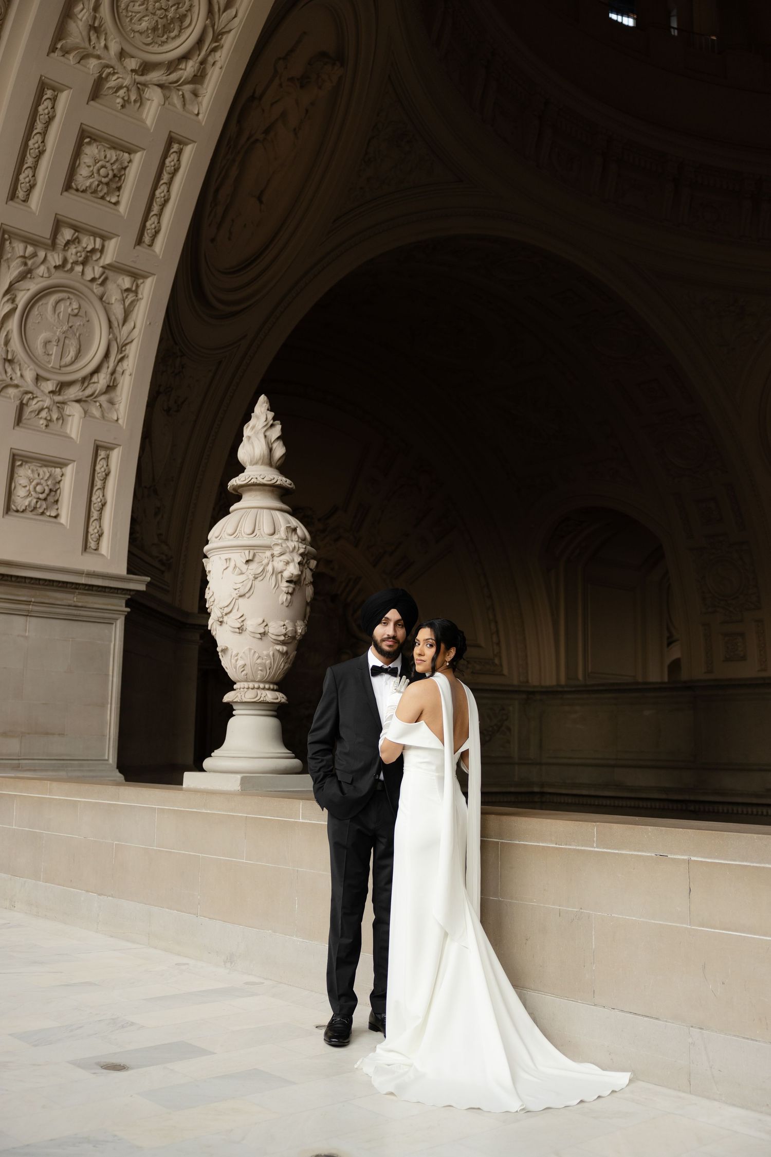 Elegant couple poses in formal wedding attire beneath ornate stone archway with decorative urn.