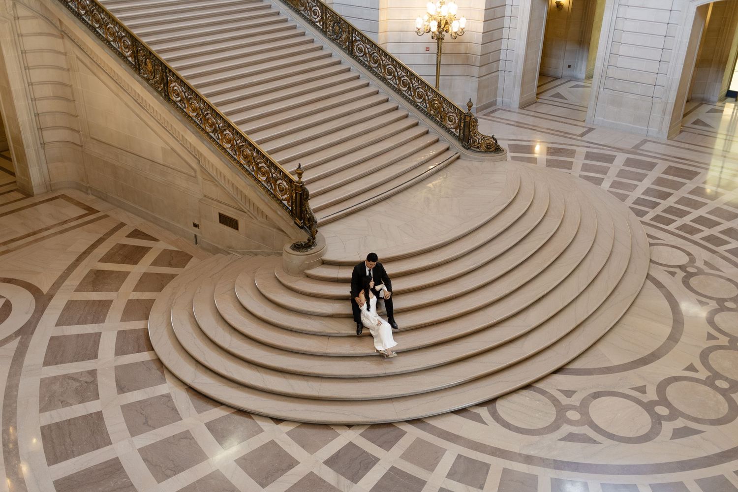 A couple descends a grand marble staircase in San Francisco City Hall with intricate floor patterns below.