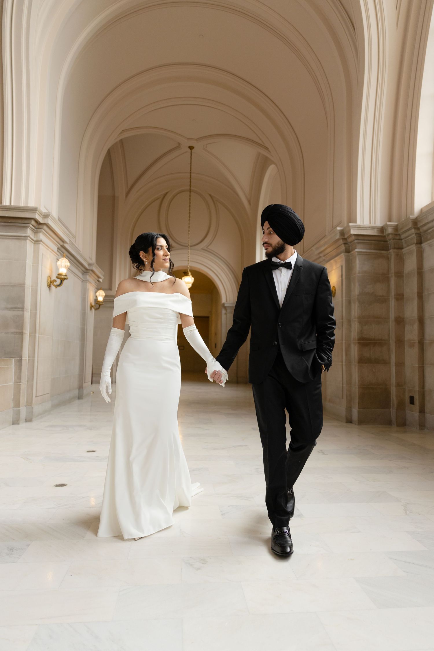 A couple in wedding attire walks through an elegant arched hallway with white marble columns and walls.