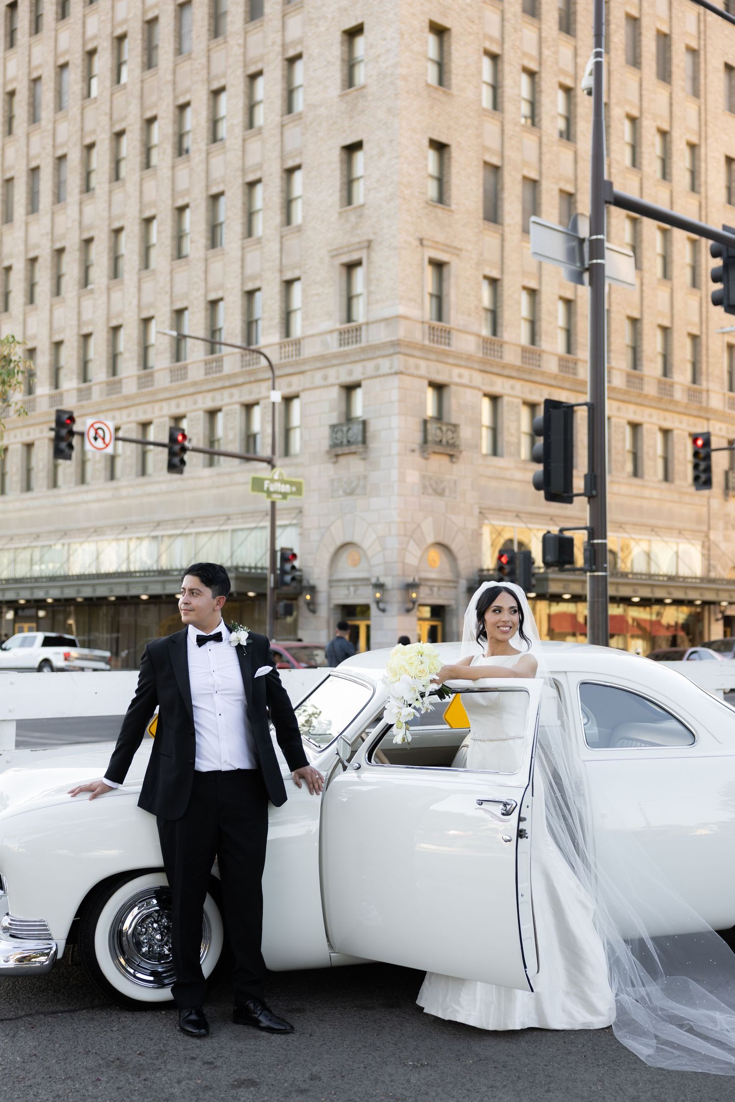 Classic white vintage car adorned with flowers serves as elegant wedding transportation on city street.