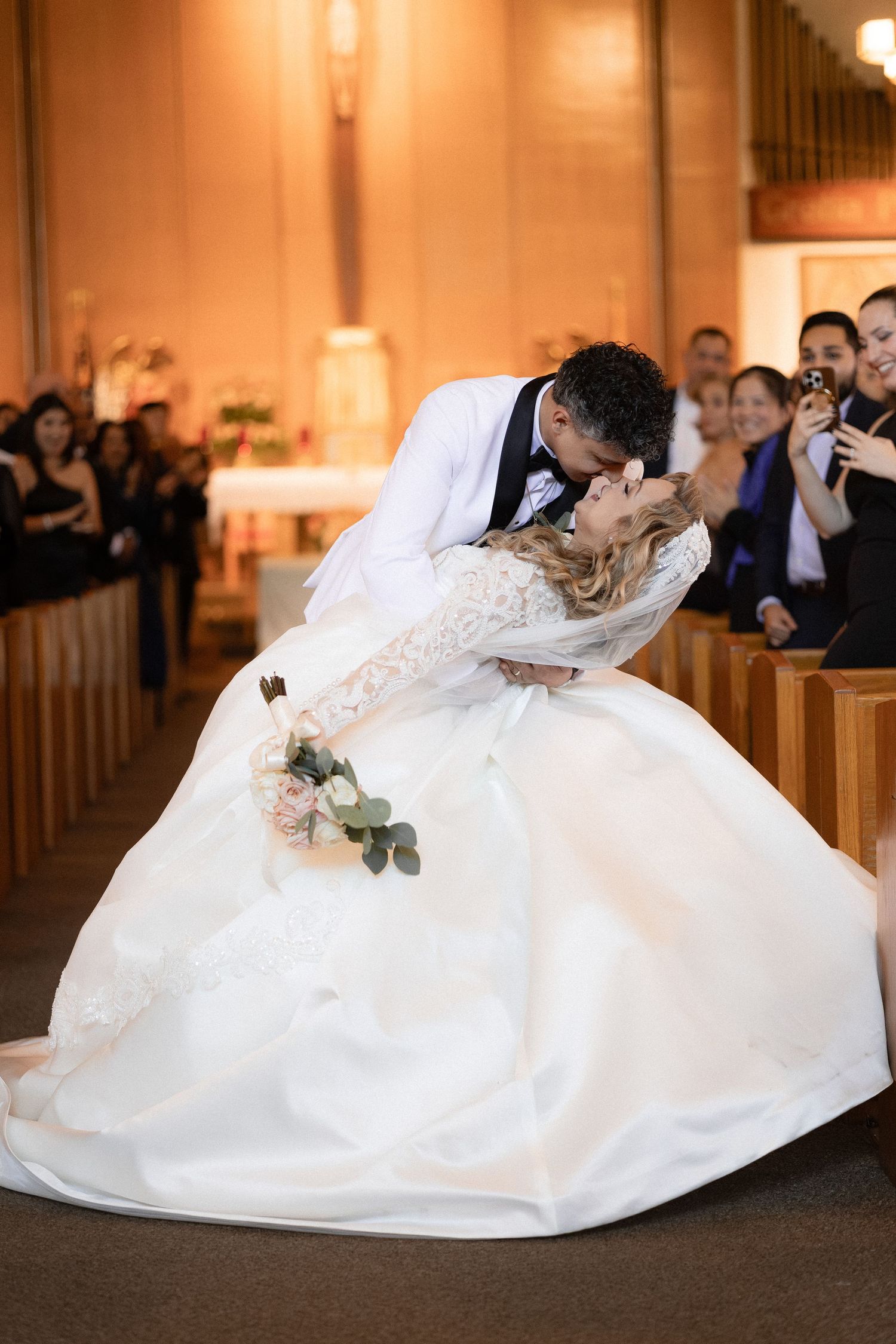 Bride in a flowing white gown shares a dramatic kiss with groom in church aisle.