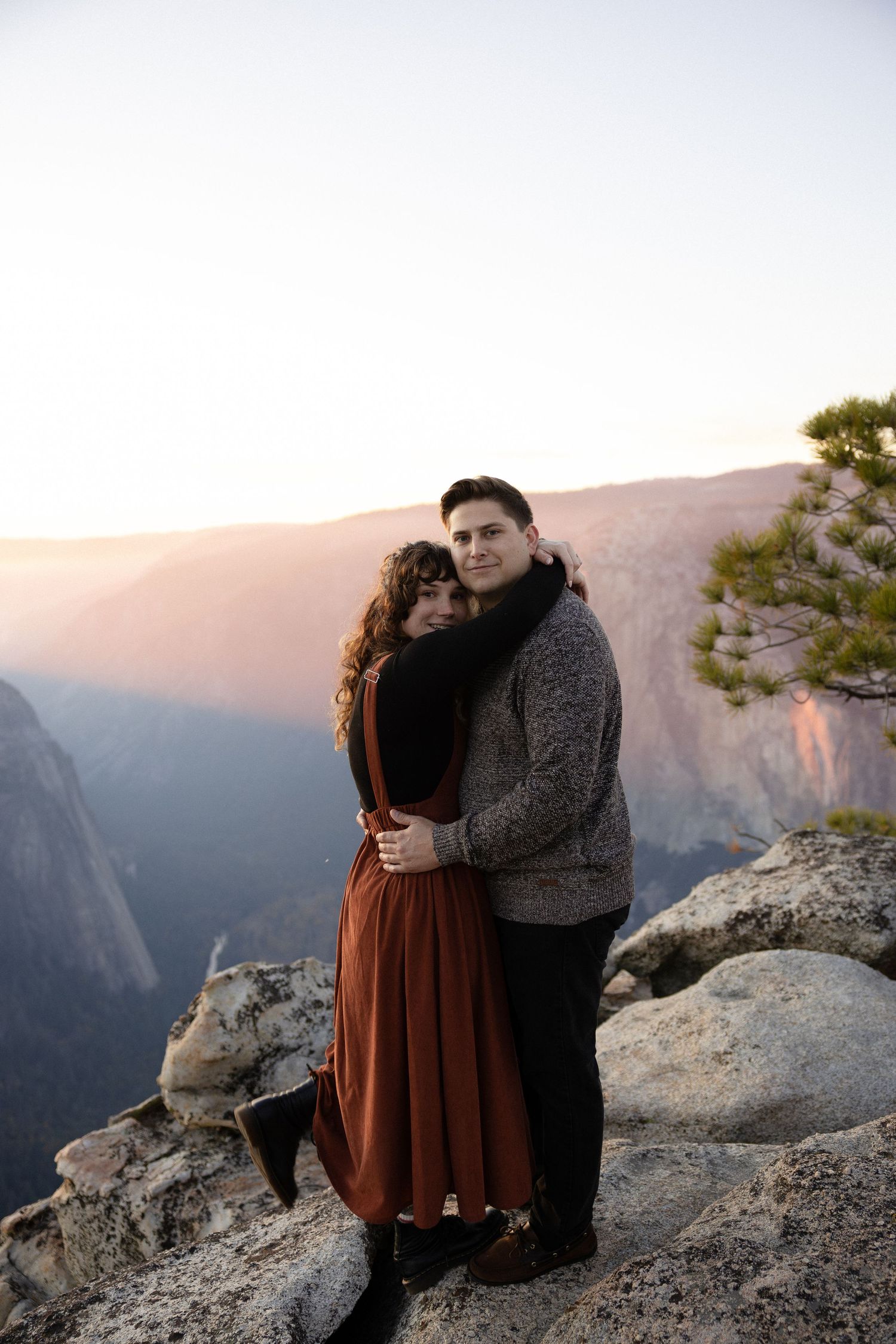 Two people share an intimate moment on a rocky cliff overlooking a vast mountain valley at dusk.