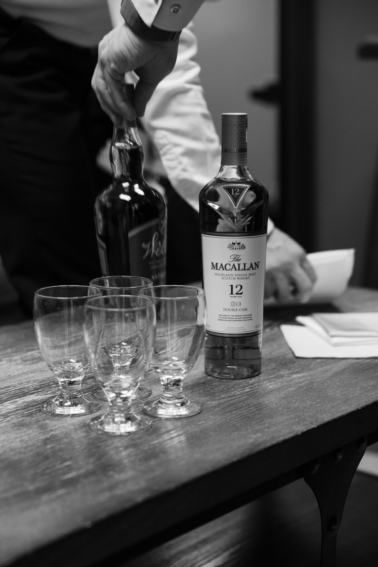 Black and white photo of Macallan 12 whisky bottle with crystal glasses on wooden table at wedding reception.