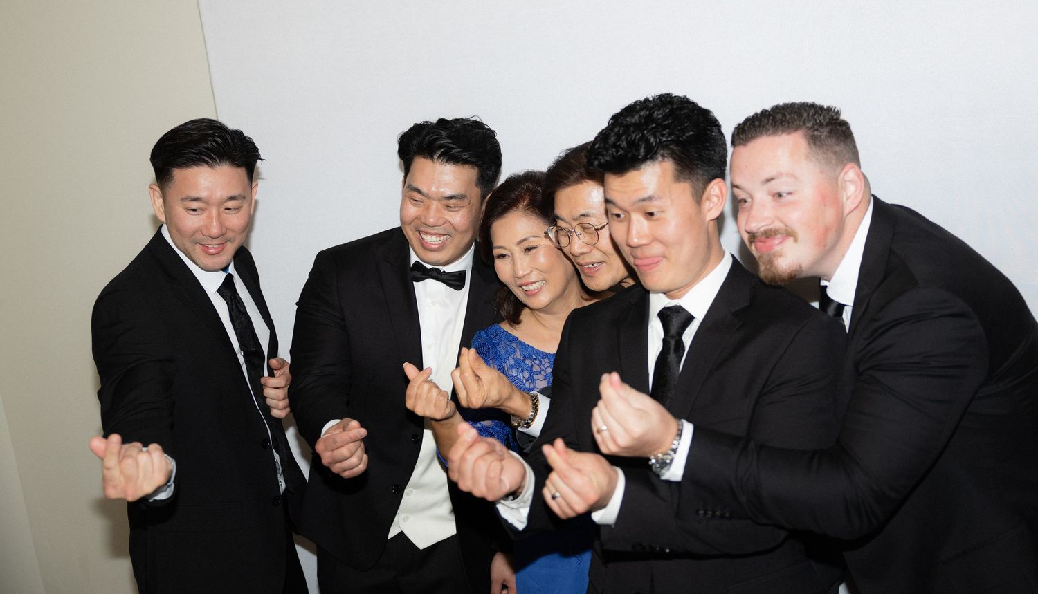 Group of six guests in formal attire pose playfully together at wedding reception against white wall.