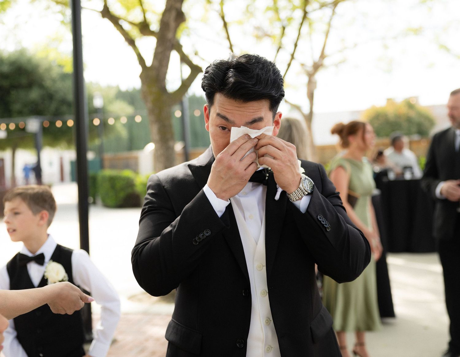 Emotional groom in black tuxedo wiping tears with handkerchief during wedding ceremony.