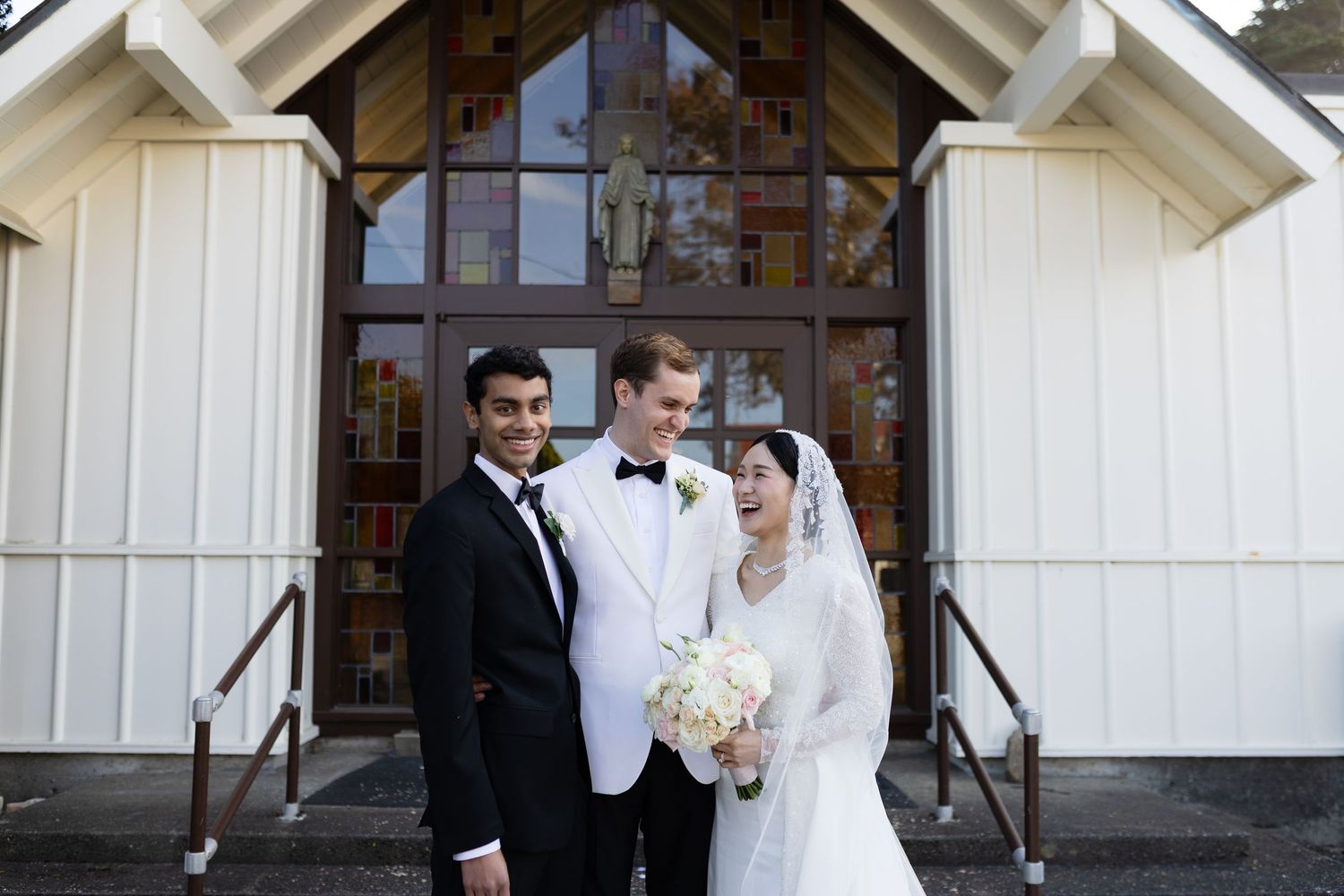 Bride and groom in white wedding attire pose with a guest outside a white chapel with stained glass windows.