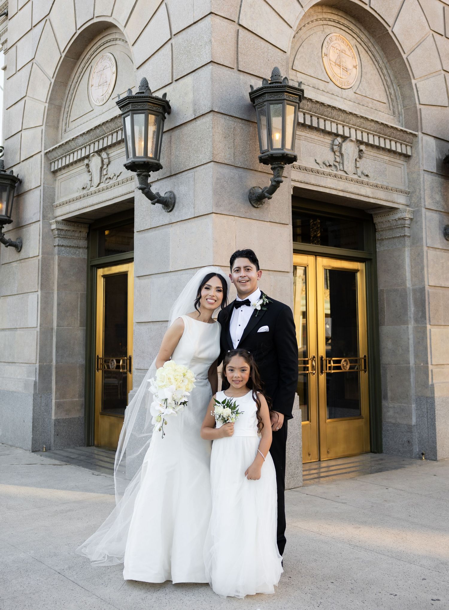 Wedding photo of couple with flower girl outside historic stone building with ornate lanterns.