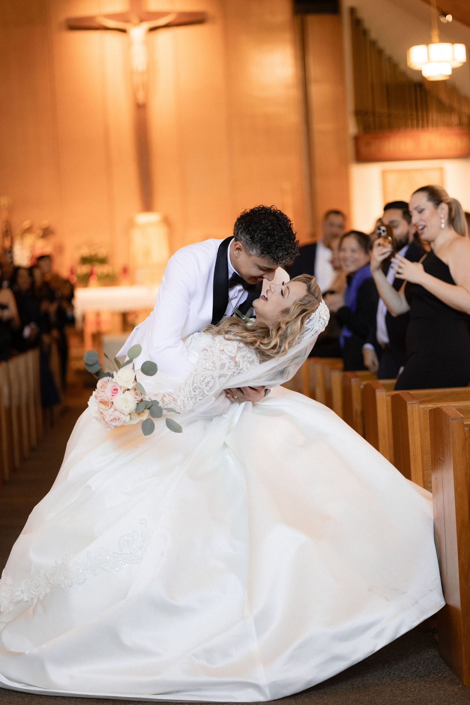 A romantic dip kiss between newlyweds in a church with a crucifix in the background.