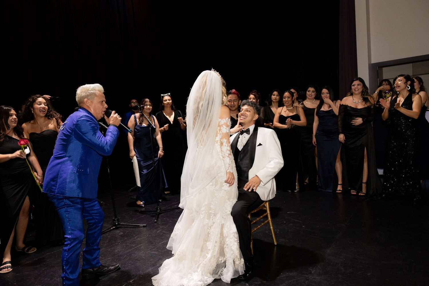 A bride in a lace gown dances with someone in a white jacket while guests watch and celebrate.