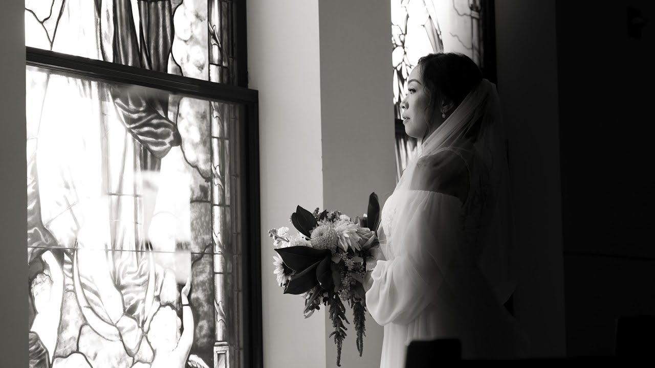 A figure in white stands near a stained glass window holding flowers in monochrome.