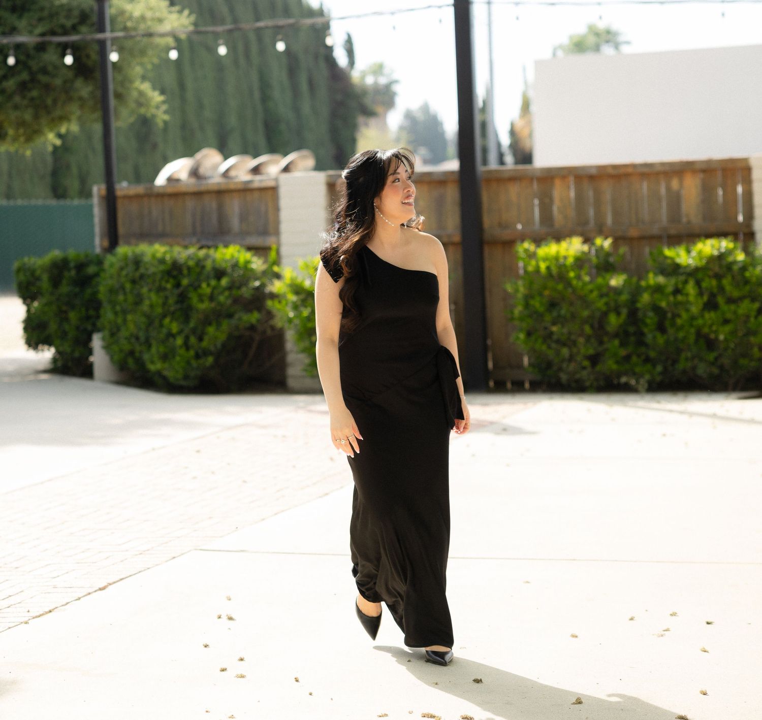 Woman in black one-shoulder jumpsuit walking outdoors at wedding venue.