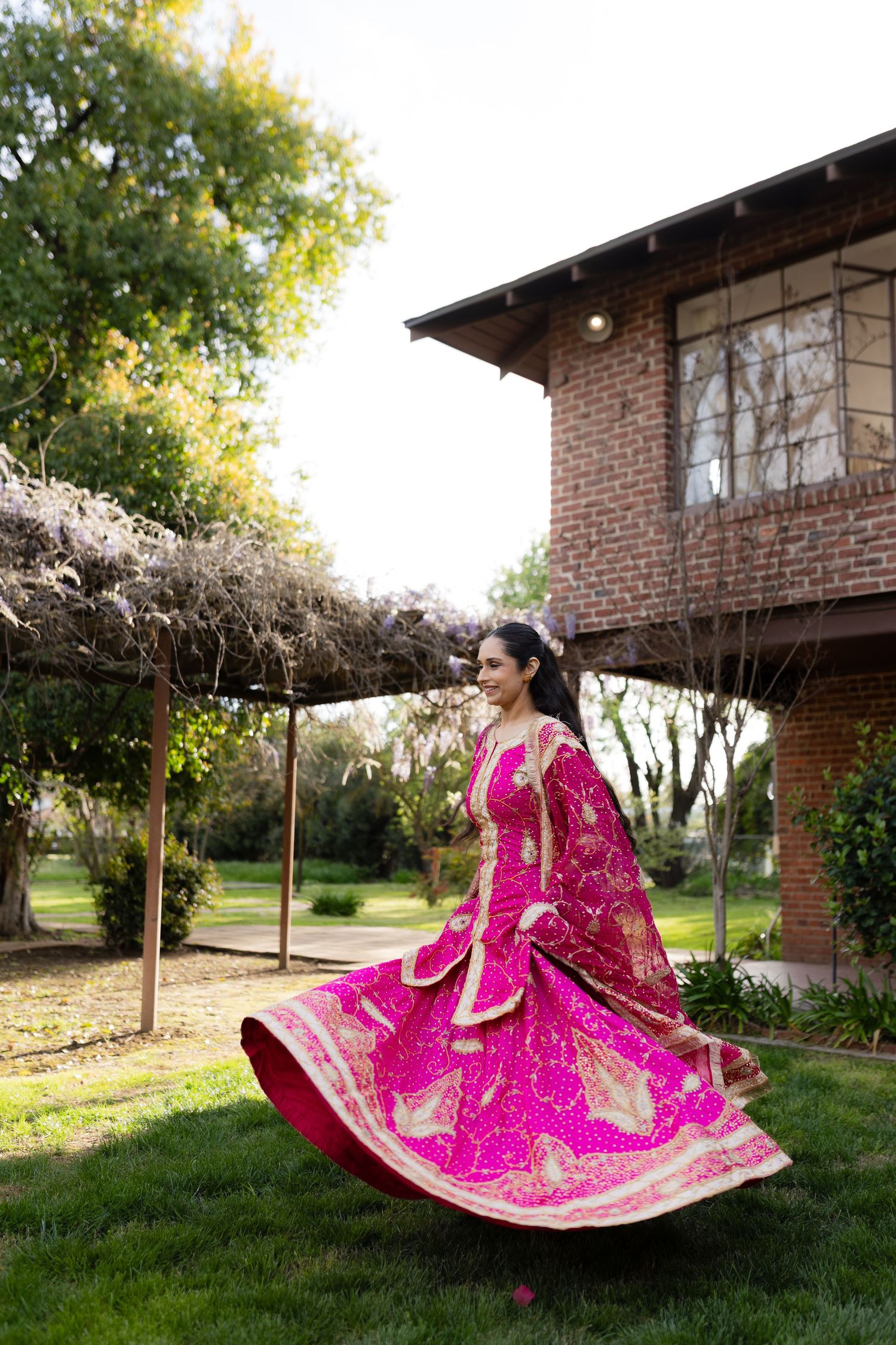 A person in a flowing pink traditional dress twirls gracefully in a garden setting with brick buildings in the background.