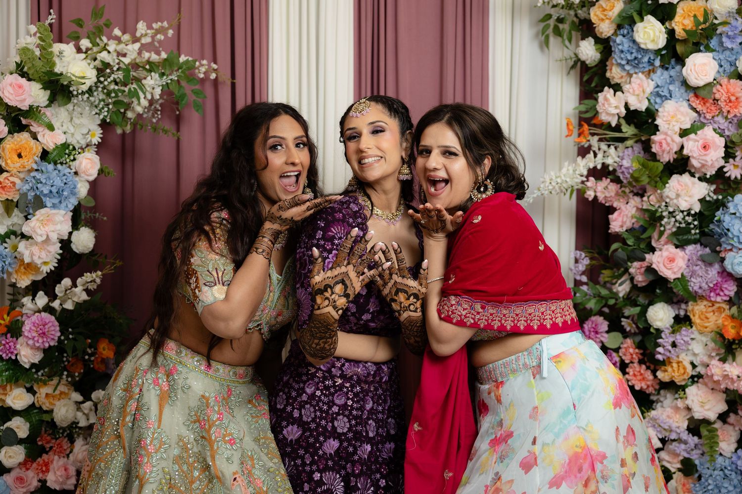 A joyful celebration with women in colorful ethnic attire laughing together by a flower wall.