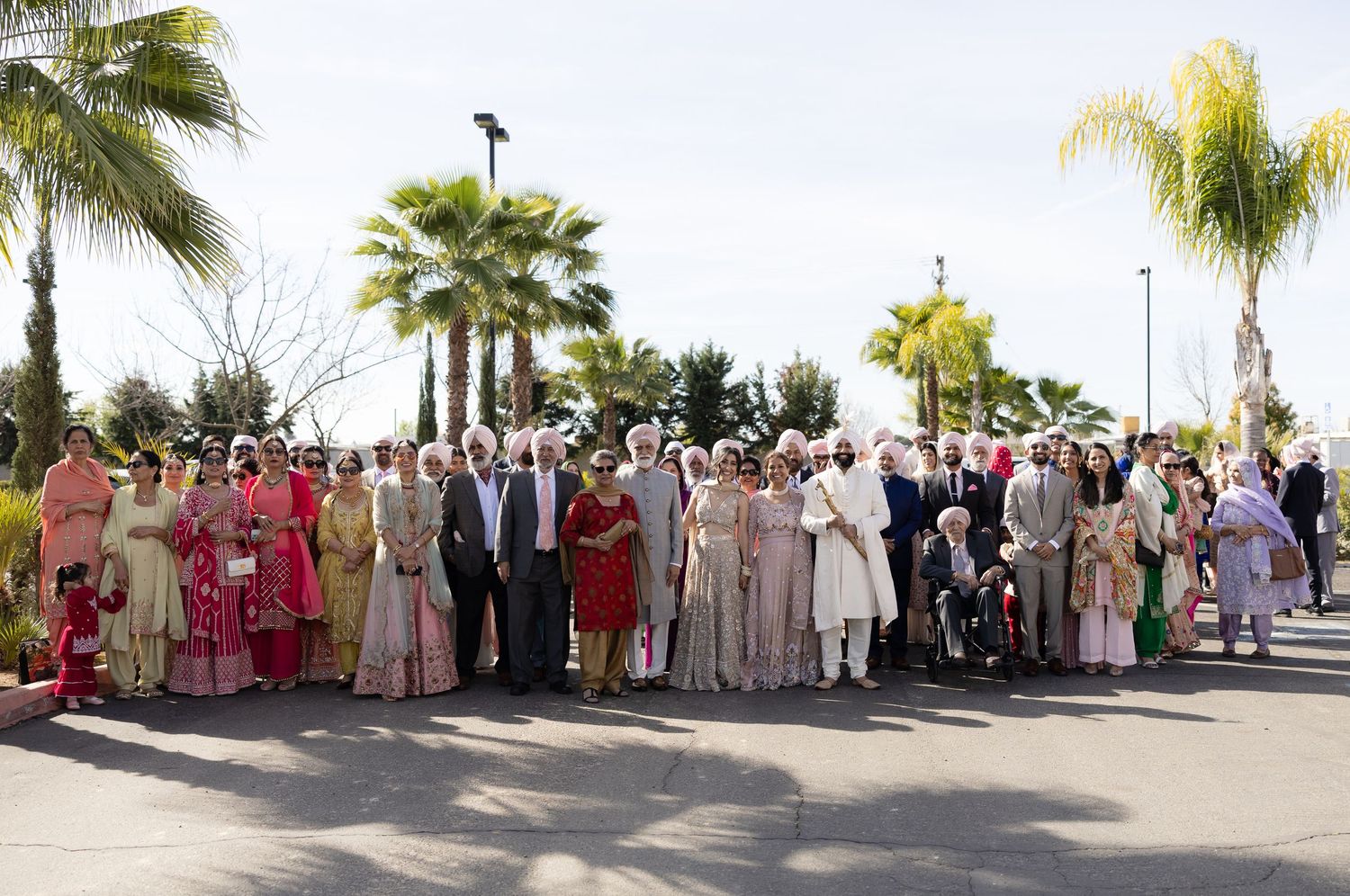 Large group of wedding guests in traditional Indian and Sikh attire pose together outdoors near palm trees.