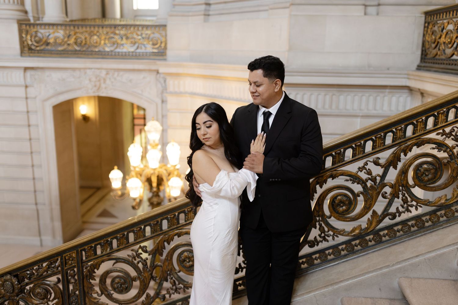 A couple shares an intimate moment on an elegant marble staircase with ornate gold railings in a grand building.