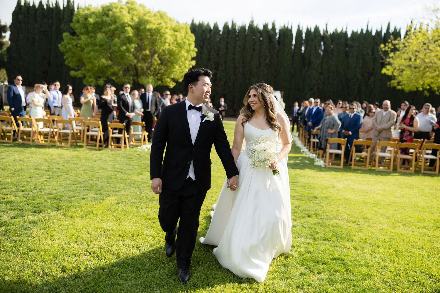 Newlyweds walk down the aisle hand in hand at an outdoor wedding ceremony with guests seated on wooden chairs.