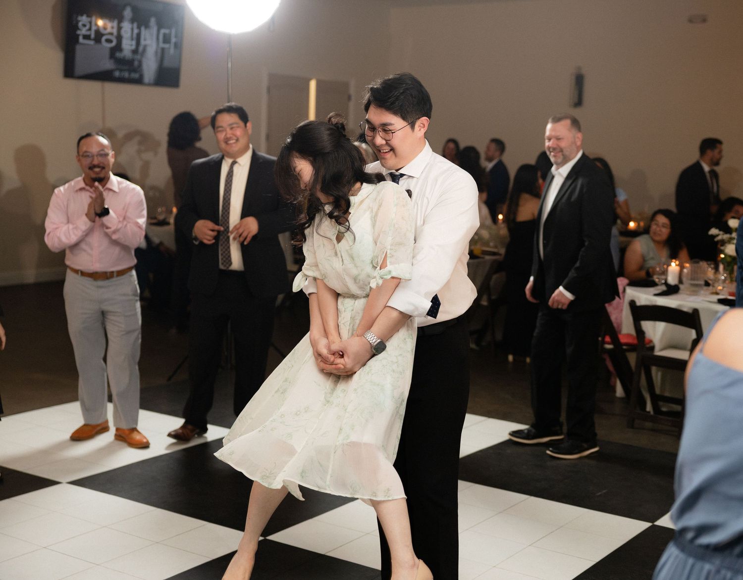 Bride and groom share first dance on black and white checkered floor surrounded by wedding guests.