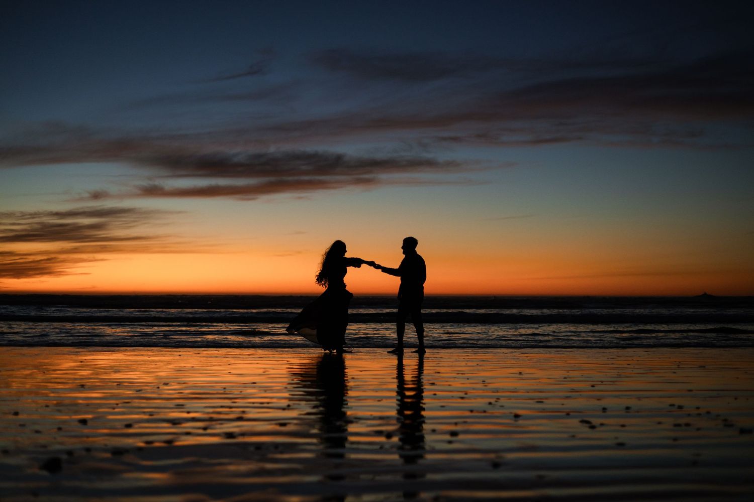 Two silhouettes stand together on a beach at sunset with a dramatic orange sky reflected in the wet sand.