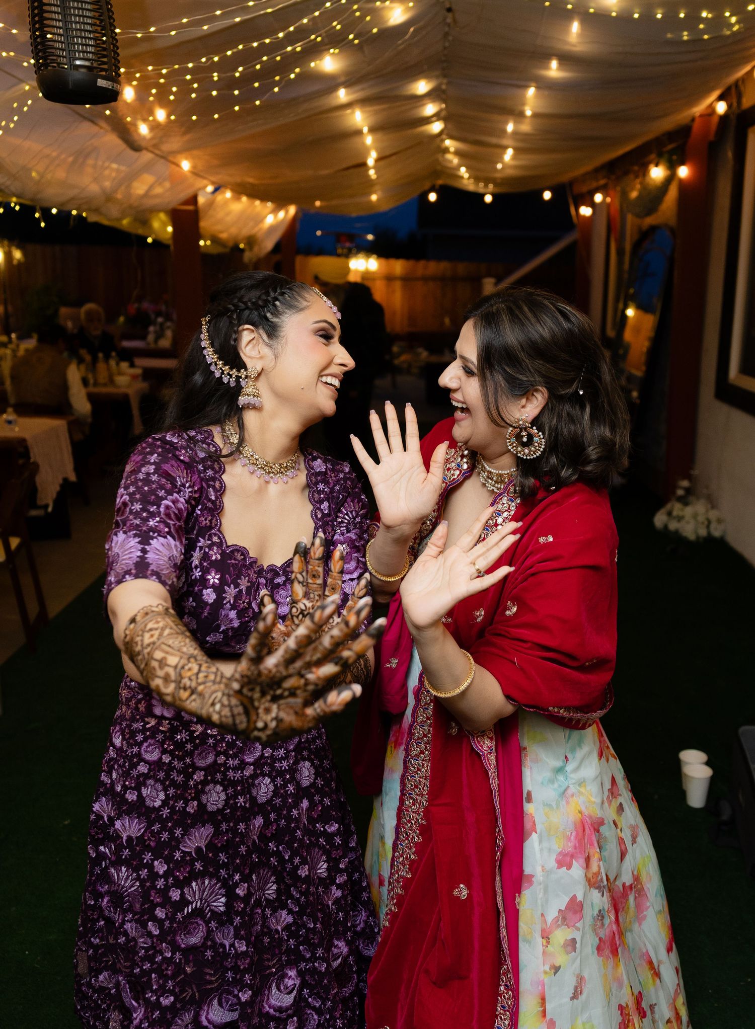 Two people in purple and red outfits share joyful laughter while dancing at a festive event with string lights overhead.