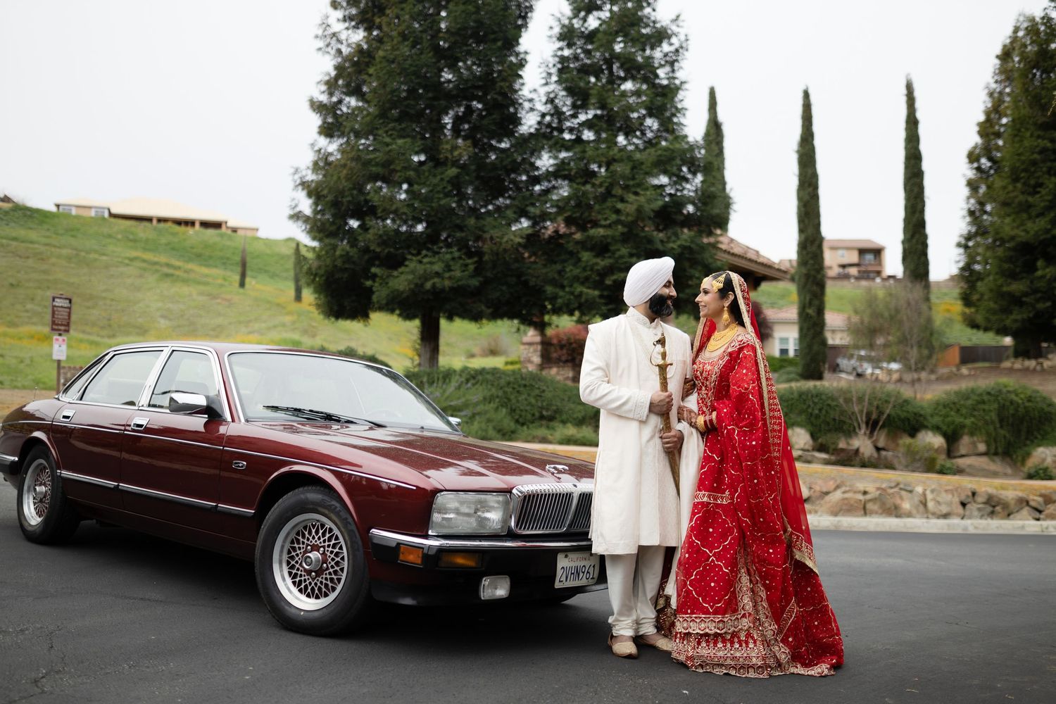 A couple in traditional South Asian wedding attire poses beside a vintage burgundy luxury car.