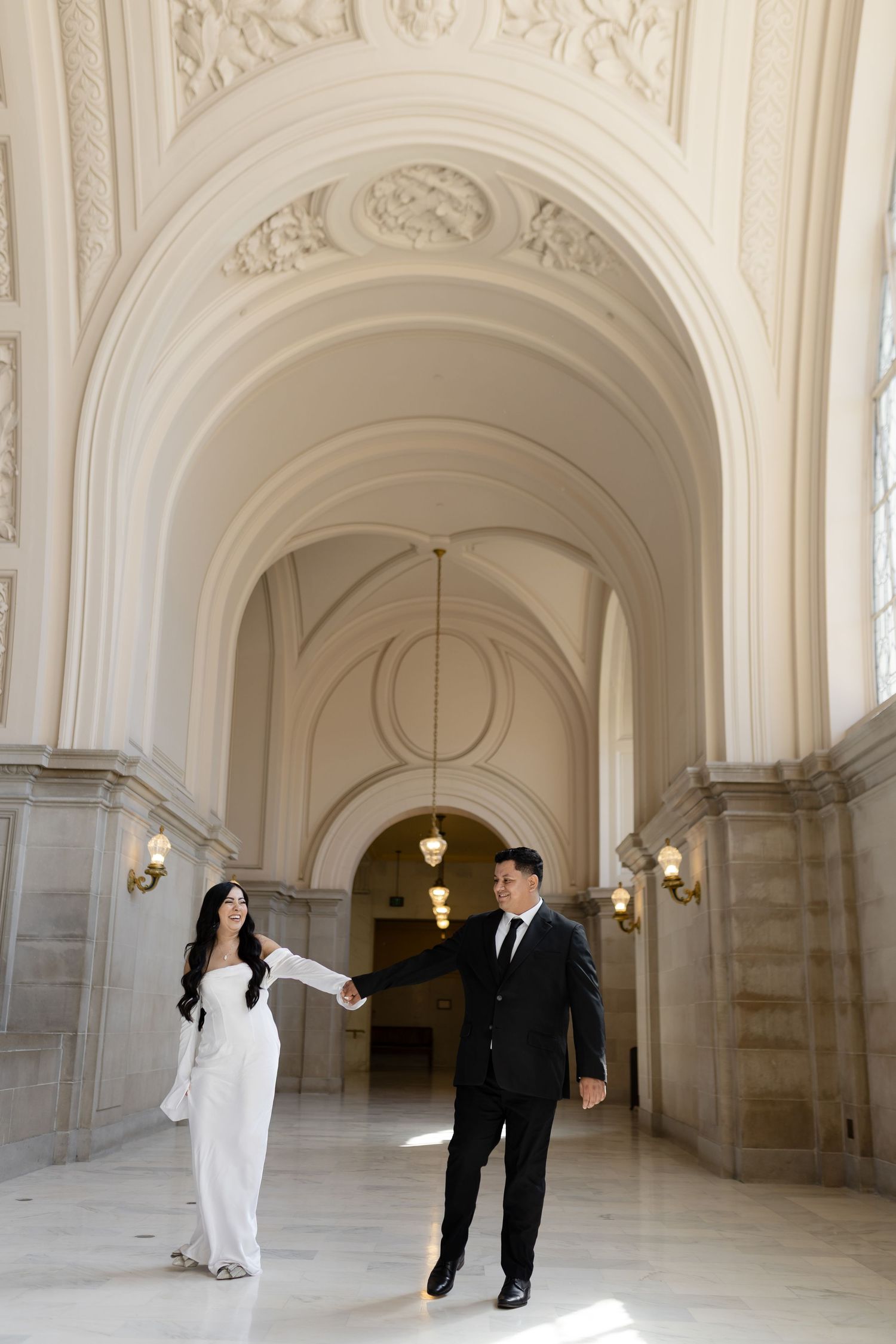 A couple in formal wedding attire dances and twirls in an elegant white marble hallway with arched ceilings and ornate details.