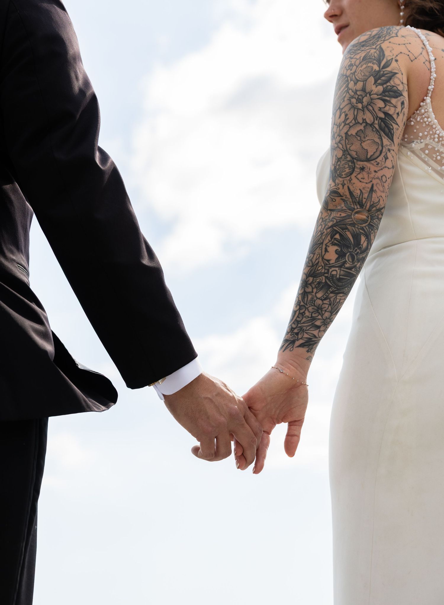 Bride with full sleeve tattoo holding hands with groom in black suit against a cloudy sky backdrop.