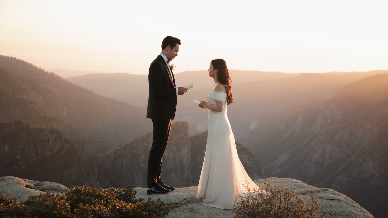 Couple exchanging vows at sunset on a rocky mountain overlook in Yosemite, bride in white gown, groom in black suit.