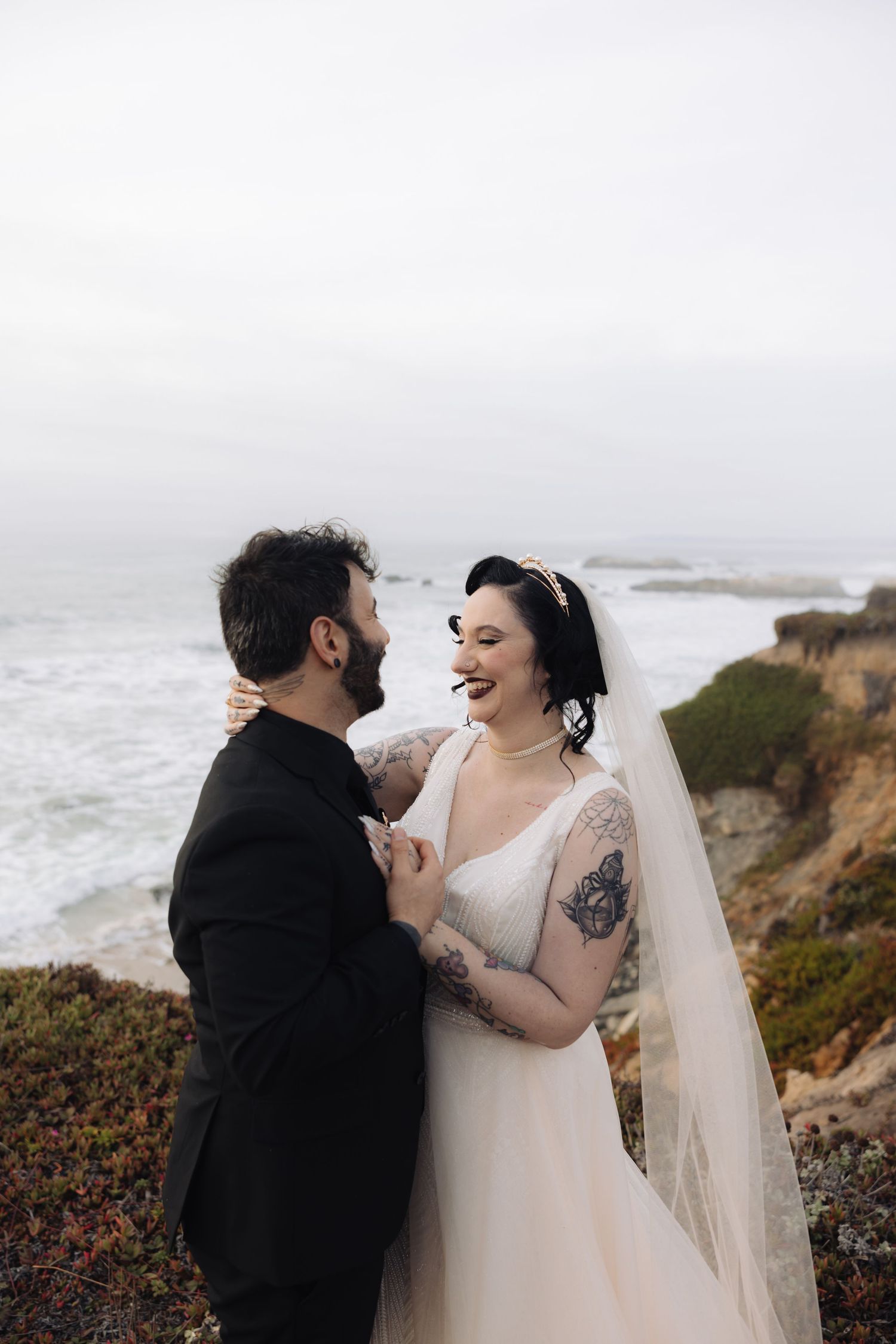 A joyful wedding couple laughs together on a misty oceanside cliff.
