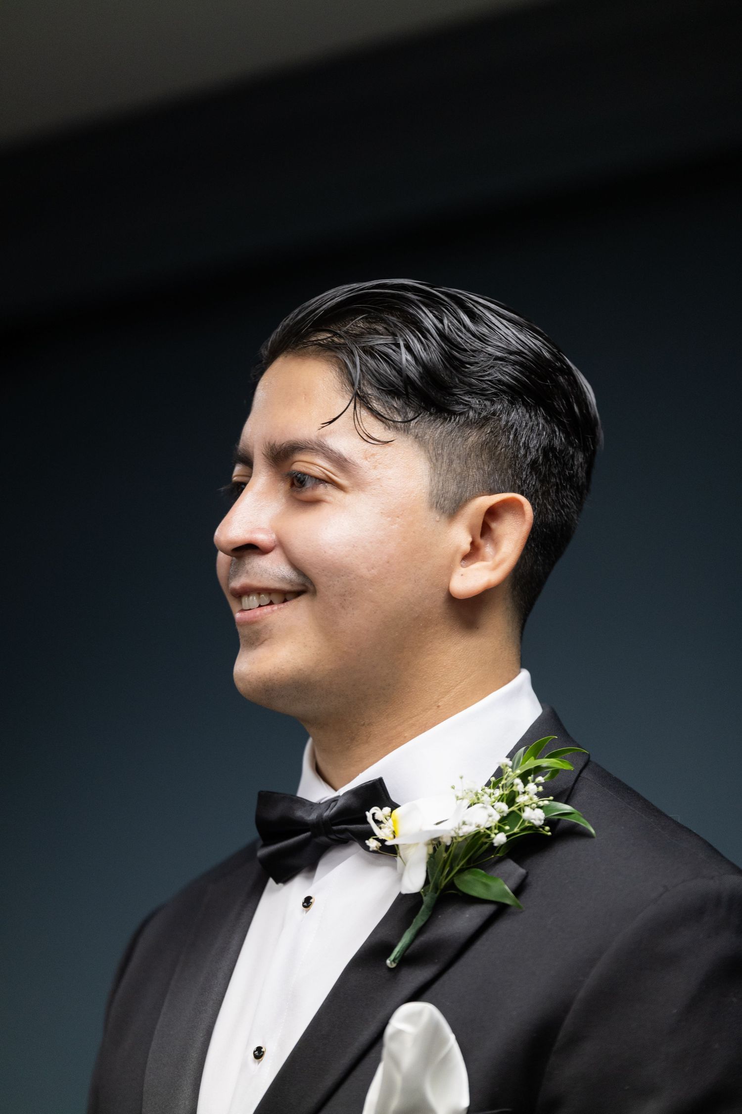 Close-up profile portrait of groom in black tuxedo with white boutonniere against dark background.