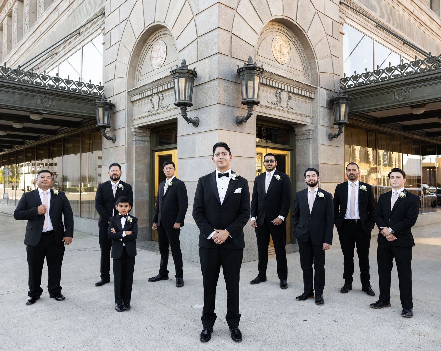 Groomsmen in black tuxedos pose for formal wedding photo outside stone building with ornate architecture.