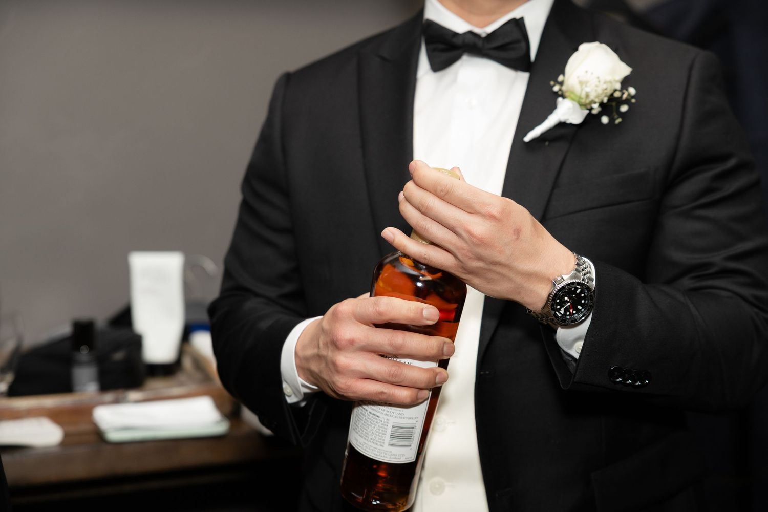 Groom in black tuxedo with boutonniere holding a whiskey bottle while wearing a luxury watch.