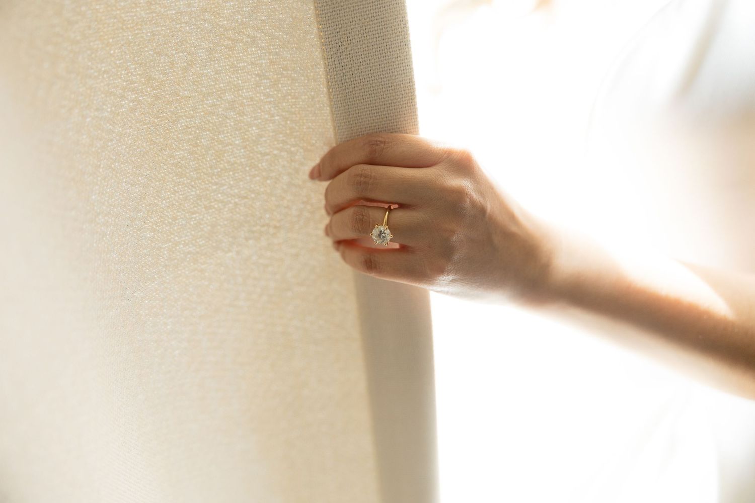Close-up of a woman's hand wearing an elegant diamond engagement ring, gently holding a white curtain.