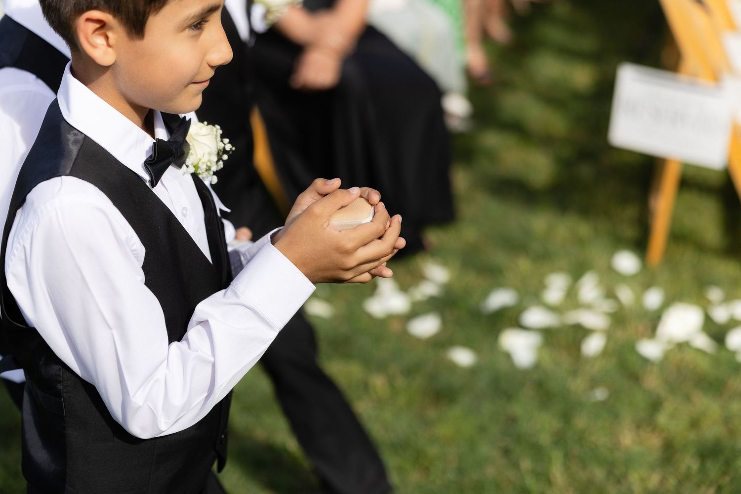 Young ring bearer in vest and boutonniere holds wedding rings at outdoor ceremony with white petals on grass.