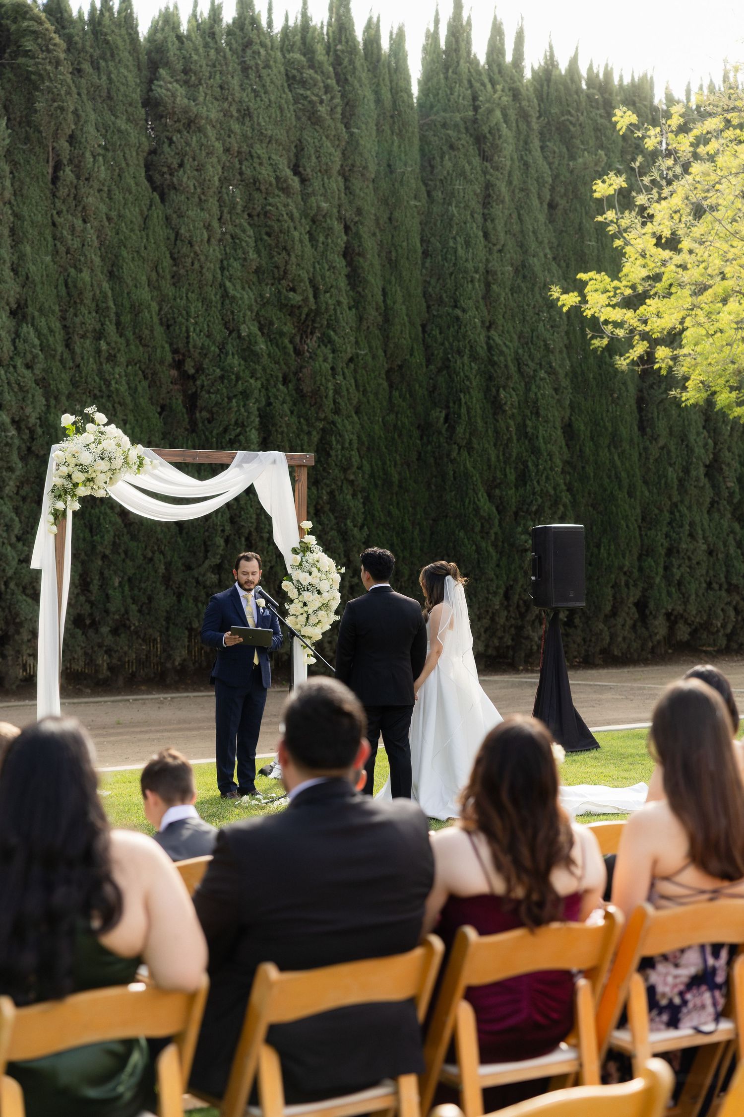 Couple exchanges vows at floral arch during outdoor garden wedding ceremony with seated guests.