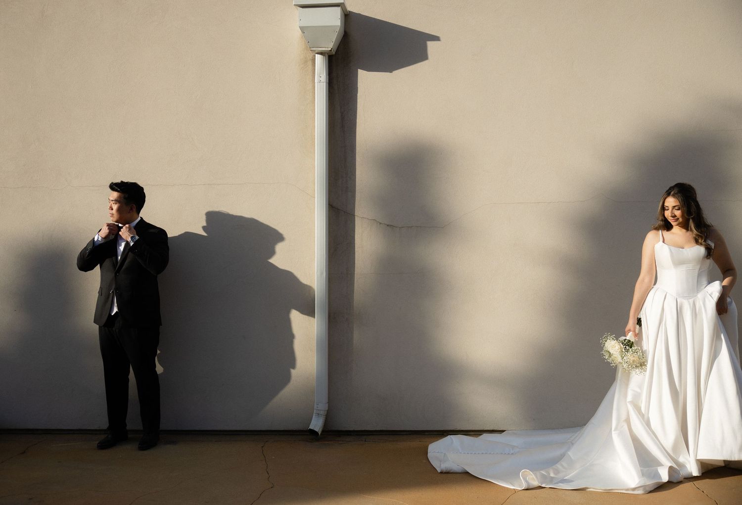 Bride in white gown holding bouquet and groom in tuxedo pose separately against sunlit wall with dramatic shadows.