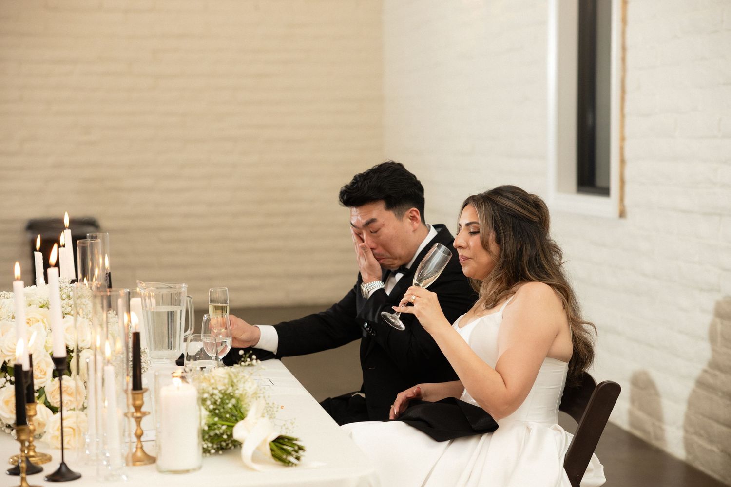 Bride and groom sit at sweetheart table with candles, sharing an emotional moment during wedding reception.