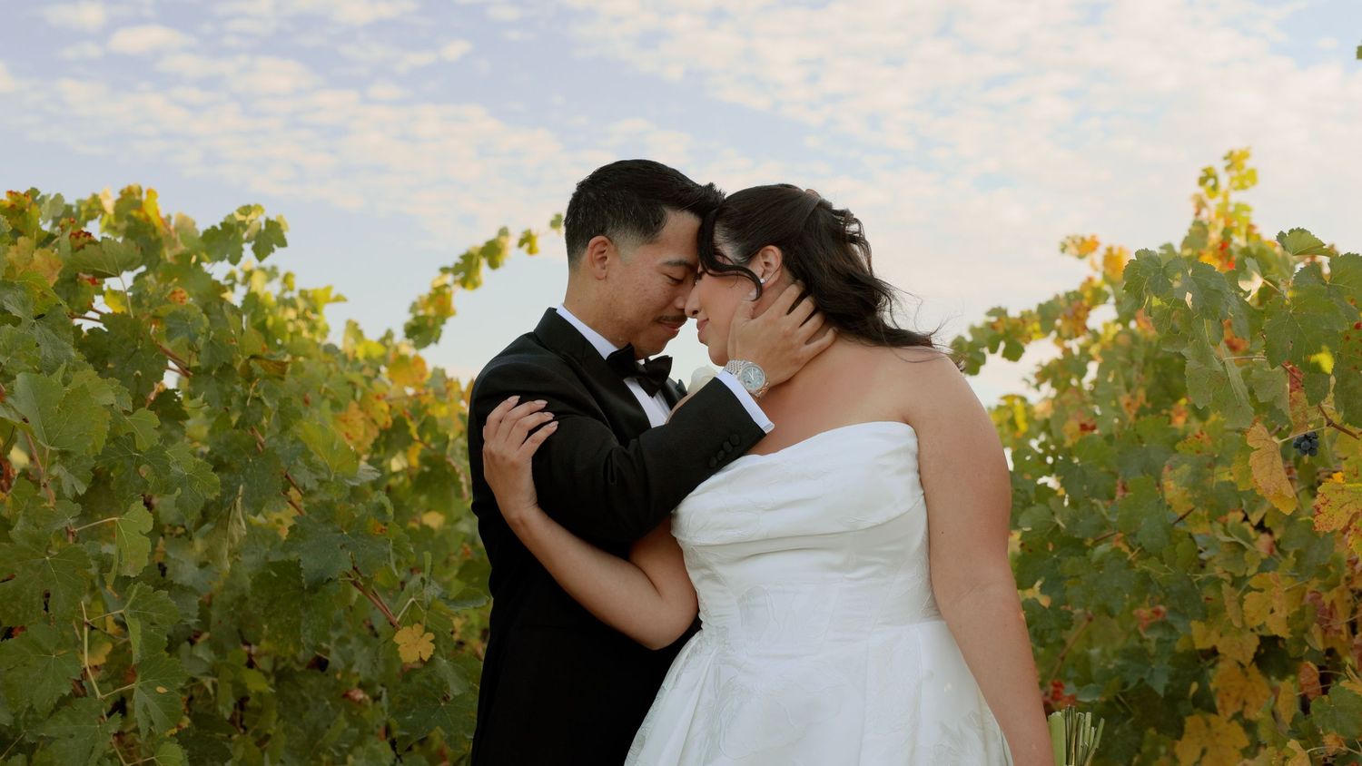 Bride and groom share a romantic moment among lush vineyard rows under a cloudy sky on their wedding day.