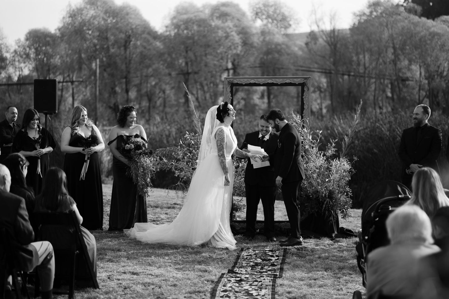 Wedding ceremony participants arranged near wooden arch structure in natural outdoor setting.
